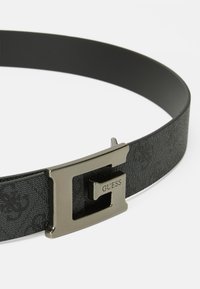 Une ceinture noire en matériau texturé avec une boucle en métal argenté poli. La boucle met en avant un 'G' prononcé et le logo "GUESS".