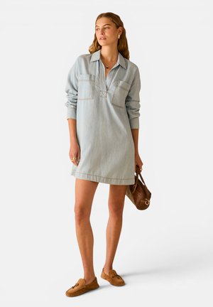 Donna che indossa un vestito di jeans azzurro chiaro con maniche lunghe e colletto, tiene una borsa marrone e indossa mocassini marroni.