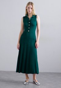 Vrouw in een mouwloze, donkergroene midi-jurk met knopen en een kraag, met zilveren platte schoenen, staand tegen een egale muur.