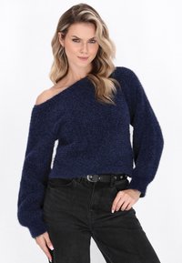 Tengerészkék, váll nélküli kötött pulóver, texturált, bolyhos felülettel, széles ujakkal és rövidített fazonnal, fekete farmernadrággal és övvel párosítva.