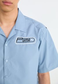 Lys blå kortermet skjorte med knappelukking, som har en svart-hvitt Puma-logo-patch på venstre bryst og en glatt tekstur.