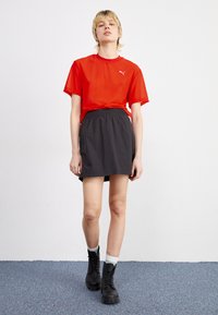 Röd genomskinlig t-shirt med kort ärm och Puma-logotyp, och matchad med en svart midjeskirts i elastiskt material; modellen bär svarta snörstövlar och grå strumpor.