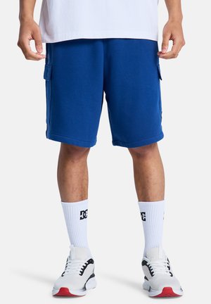 Niebieskie szorty cargo z mieszanki bawełny z bocznymi kieszeniami, w połączeniu z białymi skarpetkami z czarnym logo oraz białymi trampkami z czerwonymi akcentami.