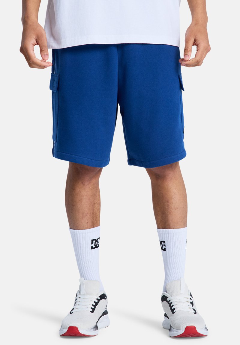 Shorts cargo bleu en mélange de coton avec poches latérales, associés à des chaussettes blanches avec un logo noir et des baskets blanches avec des accents rouges.
