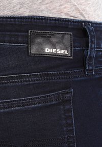 Detailní záběr tmavě modrých džínsů s viditelnou zadní kapsou a černou koženou nášivkou se značkou "DIESEL".