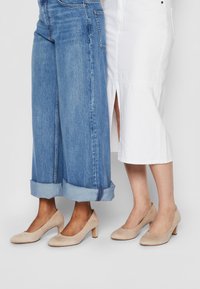 Beige semsket blokk-hæl sko, rund tå-design, brukt med oppbrettede blå jeans på den ene foten og et hvitt skjørt på den andre.