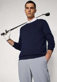 Námořnický svetr s vroubkovanými manžetami, v kombinaci se světle šedými kalhotami. Drží golfovou hůl a na levé ruce má golfovou rukavici.