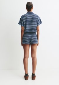 Personne avec des cheveux tressés portant un haut à rayures bleu marine et bleu clair à manches courtes et un short assorti, se tenant contre un fond blanc.