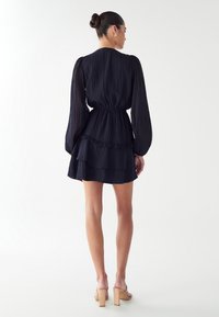 Temno modra nabrana mini obleka z dolgimi rokavi, zategnjeno pasom in nabranim robom; obuta v prozorne pete. Gladka tekstura tkanine.