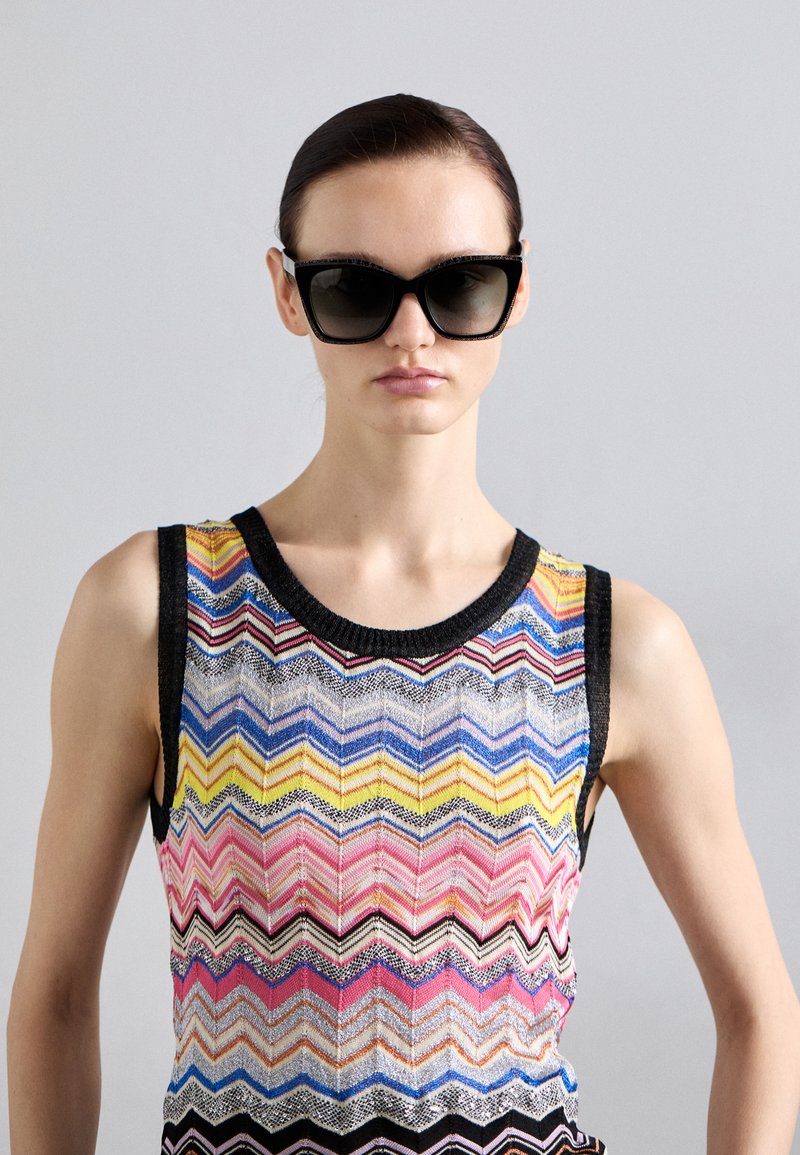 Okulary przeciwsłoneczne z wyrazistą czarną oprawą i gradientowymi soczewkami, zestawione z wielokolorowym, dzianinowym topem na ramiączkach w zygzakowy wzór z czarnymi wykończeniami.