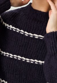 Pull tricoté bleu marine avec un col montant, présentant des rayures tressées blanches et une texture épaisse. Idéal pour des superpositions chaudes.