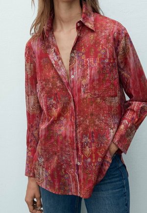 Femme portant une chemise rouge ample à motifs, boutonnée, une main dans la poche d'un jean bleu, sur fond blanc uni.