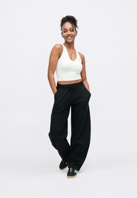 Jonge vrouw die lacht, draagt een wit mouwloos cropped topje, losse zwarte broek en zwarte sneakers, staat met handen in de zakken.