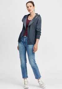 Marineblå hettegenser med glidelås og snor, kombinert med en burgunder tanktopp og lyseblå jeans, avsluttet med beige joggesko.