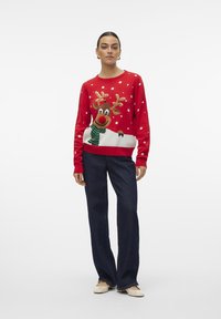 Rød strikket genser med et reinsdyrdesign, grønn skjerf og snødetaljer, kombinert med mørkeblå jeans og beige sko.