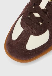 Közelről barna szarvasbőr és fehér bőr tornacipő orr rész barna fűzőkkel és texturált gumitalppal, fehér háttéren.