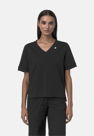 Donna con lunghi capelli scuri che indossa una maglietta nera a maniche corte con scollo a V e pantaloni neri abbinati, in piedi davanti a uno sfondo chiaro e uniforme.