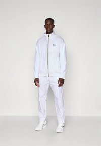 Vitt sportset i lättviktig tyg, med en jacka med dragkedja och svart logotyp, matchande byxor och vita sneakers.