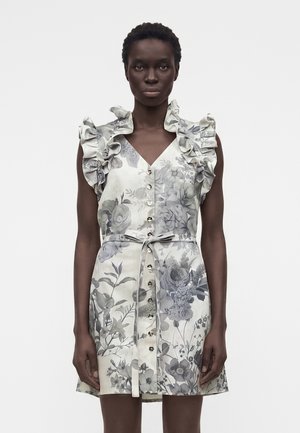 Mujer con un vestido sin mangas con botones, hombros con volantes y un estampado floral gris atado en la cintura, de pie sobre un fondo blanco.