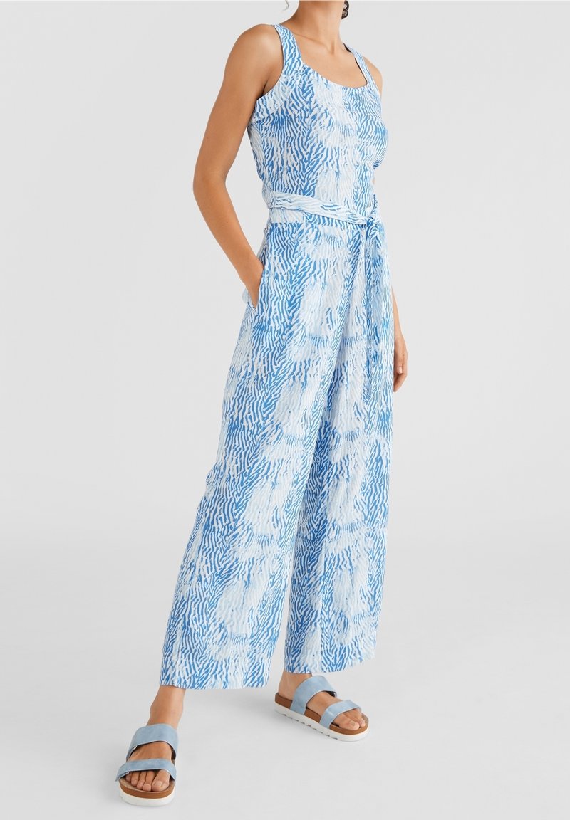 Femme portant une combinaison à jambes larges sans manches bleu et blanc à motifs avec ceinture et sandales bleu clair.