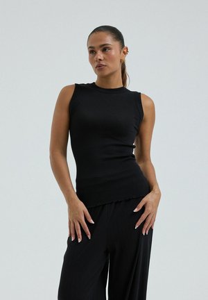 Vrouw met donker haar in een paardenstaart, gekleed in een mouwloos zwart geribd topje en zwarte broek, poserend met handen bij de heupen tegen een neutrale achtergrond.