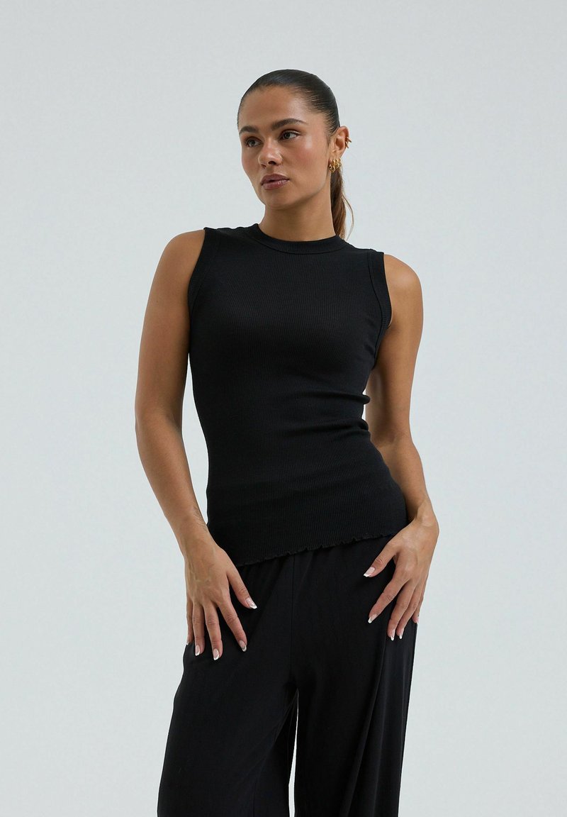 Femme aux cheveux noirs attachés en queue de cheval, portant un haut sans manches noir côtelé et un pantalon noir, posant les mains près des hanches sur un fond neutre.