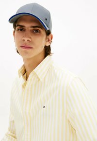 Graue Kappe mit blauen Akzenten, gelb gestreiftes Hemd mit Button-Down-Kragen und kleinem Logo. Glattes Material, lässige Passform, neutraler Hintergrund.