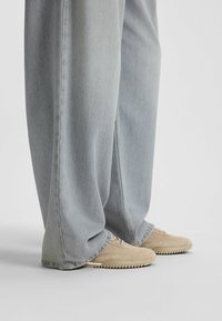 Beżowe zamszowe sneakersy z teksturowaną cholewką i gumową podeszwą, noszone z jasnoniebieskimi jeansami o szerokich nogawkach z postrzępionymi brzegami.