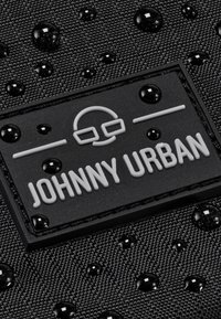Tessuto nero con un motivo testurizzato, caratterizzato da gocce d'acqua. Un'etichetta nera opaca riporta "JOHNNY URBAN" in grassetto, lettere bianche.