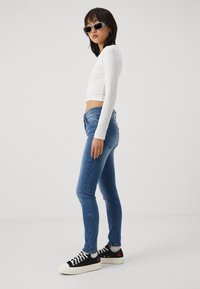 Jeans ajustados de denim azul, blusa blanca de manga larga ajustada, zapatillas negras con detalles en cereza y grandes gafas de sol grises. Textura suave.