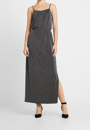 Woman wearing a sleeveless, sparkly dark gray maxi dress with a side slit and black ankle-strap heels, standing against a plain background.