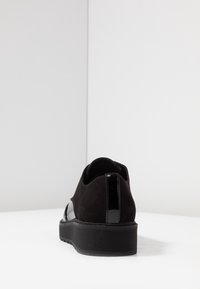 Chaussure noire avec une tige en daim, des accents en cuir verni et une semelle en caoutchouc texturée. Design lisse et épuré avec un talon subtil.