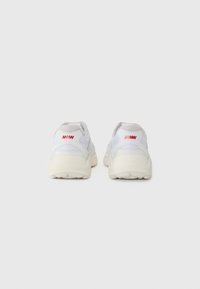 White mesh sneakers with a textured back, featuring a grey heel tab and red logo accent. Thick, curved sole for cushioning and support.