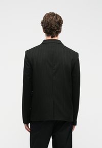 Άνδρας με σγουρά μαλλιά, φορώντας ένα μαύρο μπλέιζερ με ρίγες και μαύρο παντελόνι, που στέκεται με την πλάτη γυρισμένη σε έναν απλό λευκό φόντο.