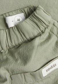 Tissu vert clair avec une texture douce, présentant des fronces à la taille et un détail de bouton. Une étiquette portant l'inscription "newbie" est visible.