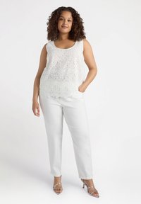 White lace tank top with a scalloped hem, paired with white straight-leg pants. Model wears silver heeled sandals, hands in pockets.