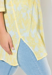 Ljusgul och blå mönstrad blus i en texturerad tyg, med en slits på sidan och vida ärmar, matchad med ljusblå jeans.