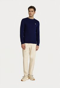 Joven de pie, vistiendo un suéter azul marino, pantalones beige y zapatillas beige, contra un fondo claro y liso.