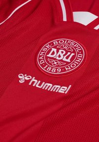 Maglia sportiva rossa con il logo della Federazione Calcistica Danese (DBU) e la scritta "Dansk Boldspil Union 1889" con il logo bianco di Hummel sotto.