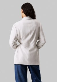 Jasnoszary marynarka o dopasowanym kroju, ze strukturalnymi ramionami i gładką fakturą, w połączeniu z niebieskimi spodniami typu wide-leg.