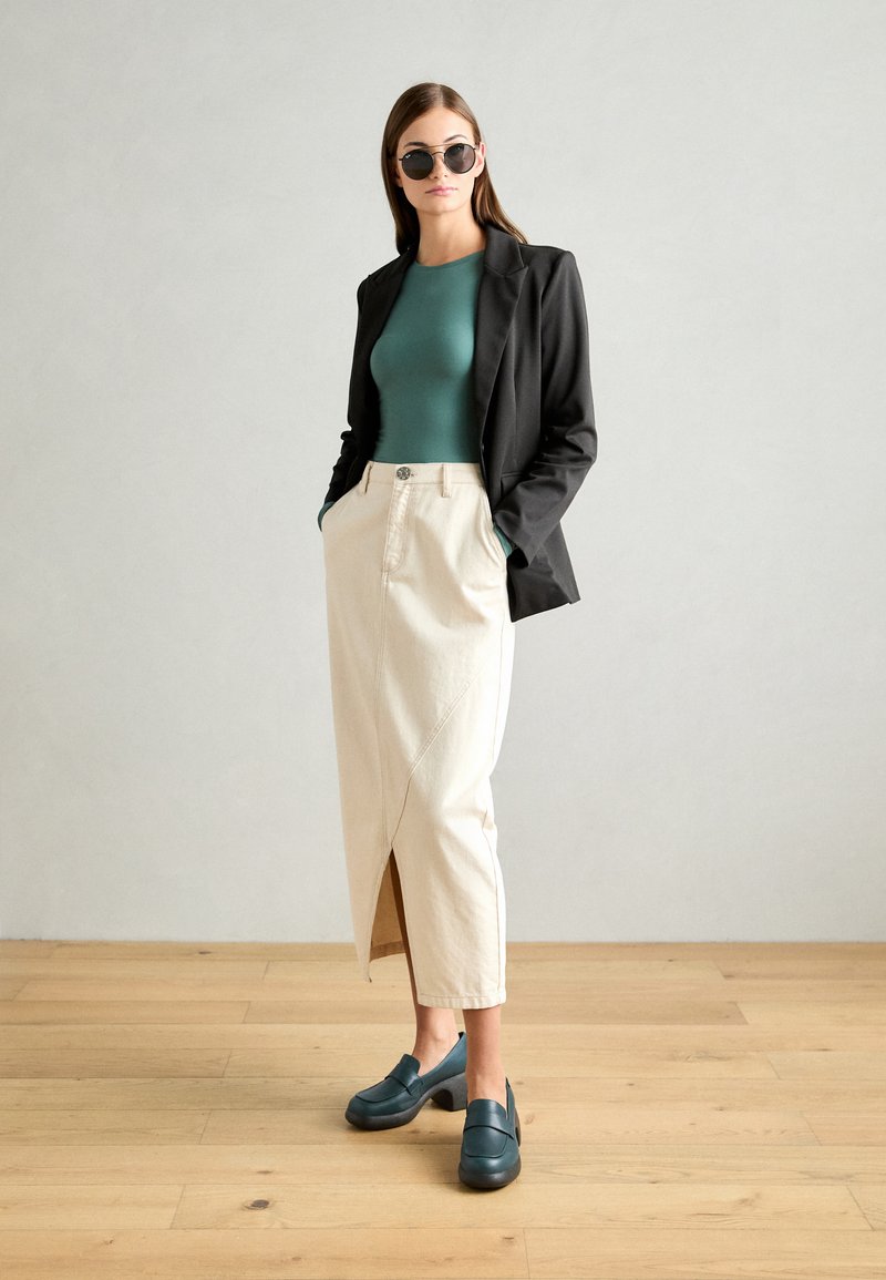 Blazer nero, top fitted teal, gonna crema con uno spacco frontale e zoccoli teal scuri. Texture lisce con un design moderno.