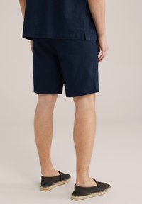 Tengerészkék pamut rövidnadrág laza szabással, oldalzsebekkel és letisztult szegéllyel. Fekete slip-on cipőkkel viselve, a lábak részben láthatók.