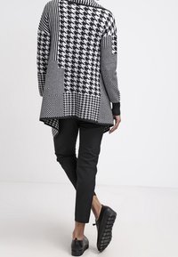 Černobílý patchwork cardigan se různými geometrickými vzory, dlouhými rukávy a asymetrickým lemem, spárovaný s černými střihovými kalhotami a botami.