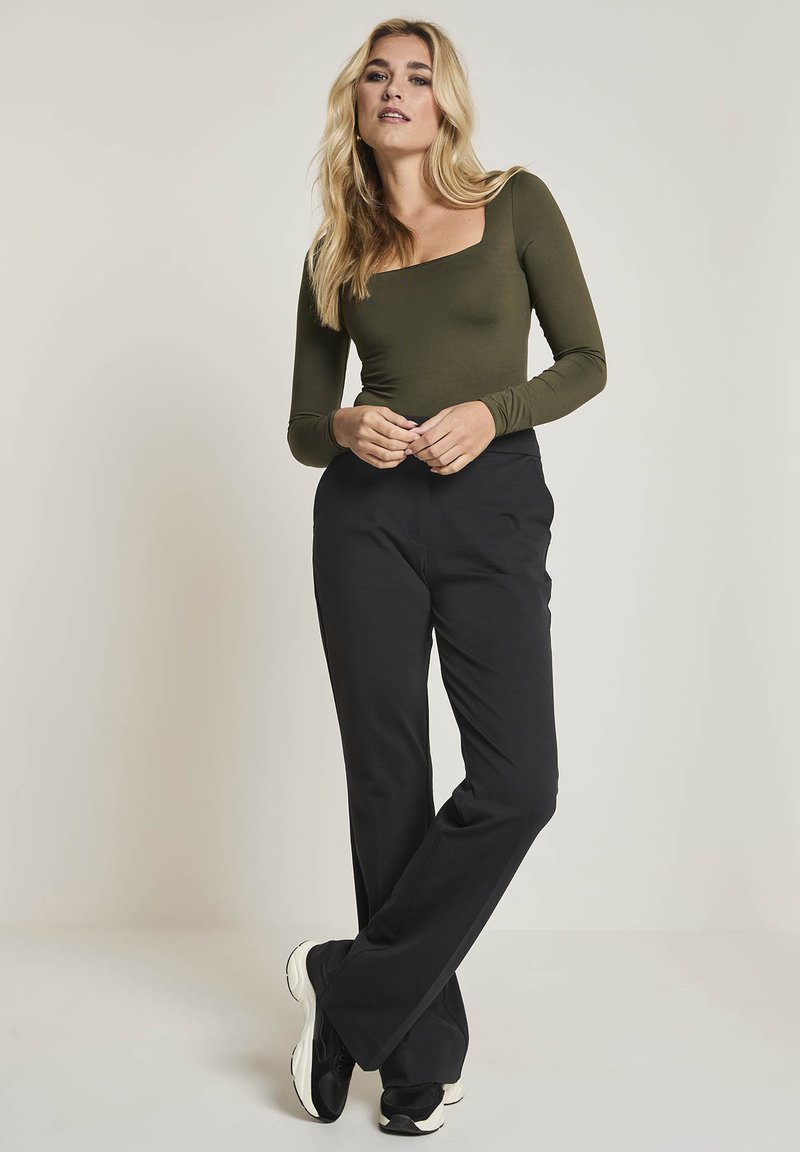 Kvinna med långt blont hår klädd i en olivgrön topp med lång ärm och fyrkantig halsringning, svarta byxor och svartvita sneakers som står med benen i kors.