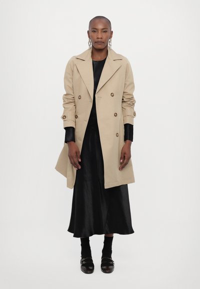 Beige double-breasted trench coat featuring large buttons, wide lapels, and rolled-up sleeves, worn over a black dress and black footwear.