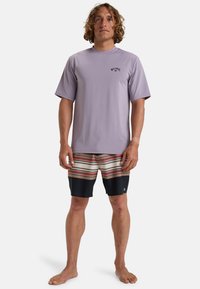 Rash guard de manga corta en un tono púrpura claro con logo negro, combinado con pantalones cortos de baño a rayas multicolores en tonos tierra, con un fondo azul oscuro.