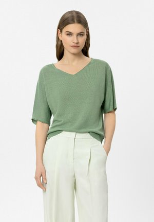 Frau mit glatten braunen Haaren trägt einen locker sitzenden grünen V-Ausschnitt-Pullover und hoch taillierte weiße Hosen mit weitem Bein, steht mit einer Hand in der Tasche.