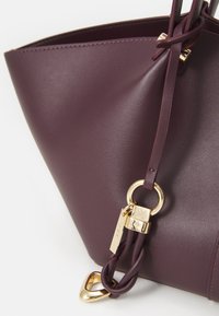 Sac seau en cuir bordeaux avec une texture lisse, relié par un anneau en or et un fermoir. Présente des accents tressés et un design épuré et moderne.