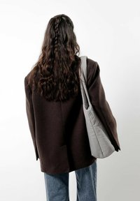 Ciemnobrązowy, dużych rozmiarów żakiet z wyraźnie zarysowanymi ramionami, zestawiony z jasnoszarym, pikowanym torbą noszoną na ramieniu, na białym tle.