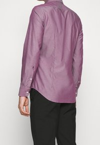 Muž v dlouhém fialovém košili s dlouhým rukávem a černých kalhotách, zobrazený zezadu na jednobarevném světlém pozadí.